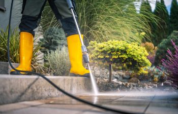 A gardener in bright yellow boots operates a pressure washer to clean a stone patio in a beautifully landscaped garden. Lush greenery and colorful plants surround the area
